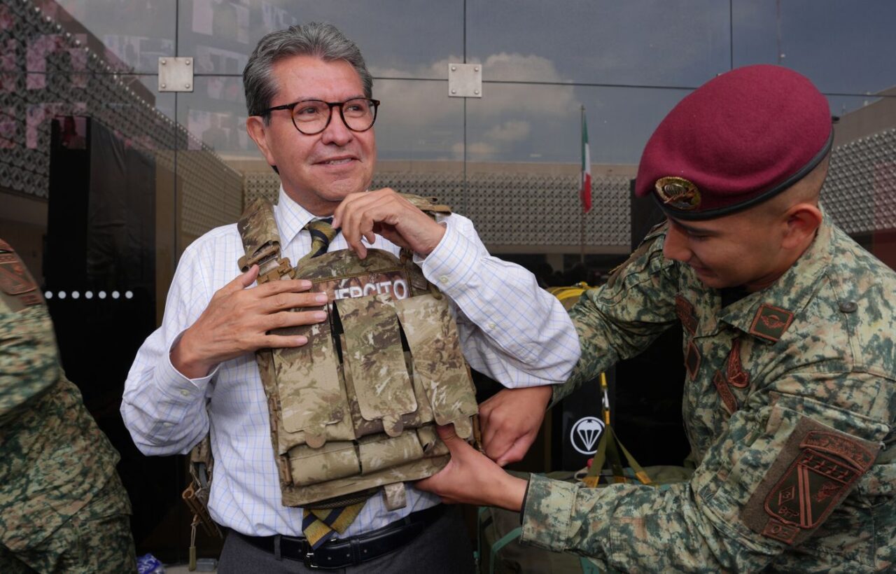 Ricardo Monreal ajusta un chaleco táctico con ayuda de un militar durante una exhibición.