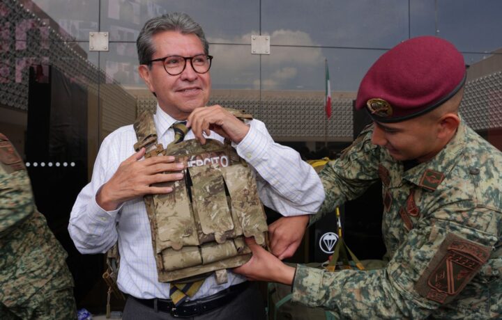 Ricardo Monreal ajusta un chaleco táctico con ayuda de un militar durante una exhibición.